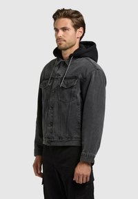 Homme portant une veste en jean noire délavée par-dessus un sweat à capuche noir, debout de profil devant un fond gris clair uni.
