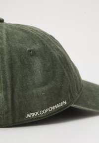 Căciulă verde din bumbac, cu o vizieră curbă și brodat "ARKK COPENHAGEN" în alb pe lateral. Textură moale, design structurat.
