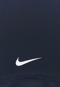Biały logo Nike w kształcie łyżwy na ciemnoniebieskim materiale z widocznym szyciem na dolnej krawędzi.