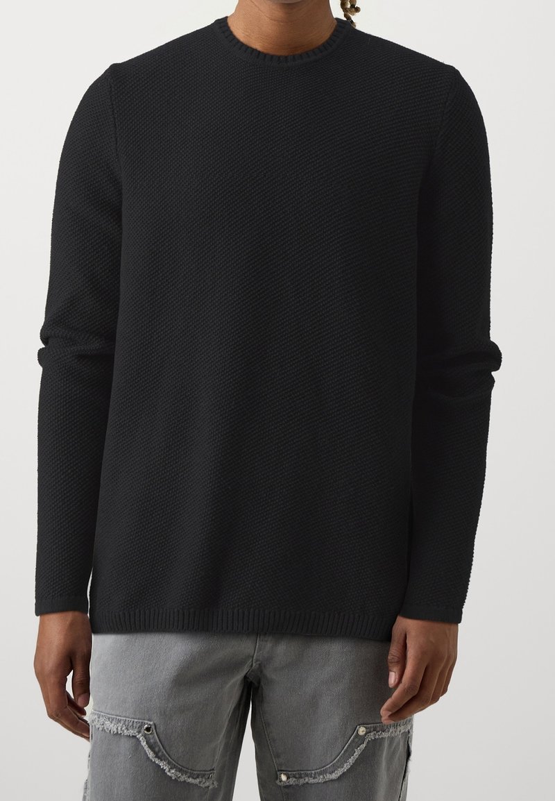 Schwarzer Strickpullover mit rundem Ausschnitt, langen Ärmeln, strukturiertem Stoff und geripptem Saum. Gepaart mit grauen, abgenutzten Jeans.