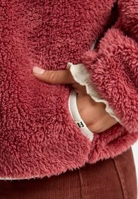 Giacca in pile di morbido materiale rosa chiaro con una consistenza soffice. Presenta una tasca con rifinitura bianca e dettagli ondulati al polsino della manica.