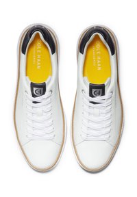 Weiße Ledersneaker mit schwarzem Fersenriegel, weißen Schnürsenkeln und einer leuchtend gelben Innensohle mit Branding. Gummisohle mit einem braunen Akzent.