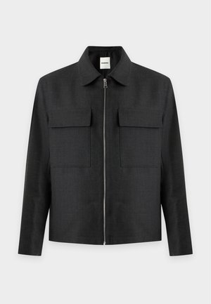 Homme portant une veste noire, une chemise rayée marron, un pantalon blanc, une ceinture noire et des chaussures noires, debout devant un fond clair uni.