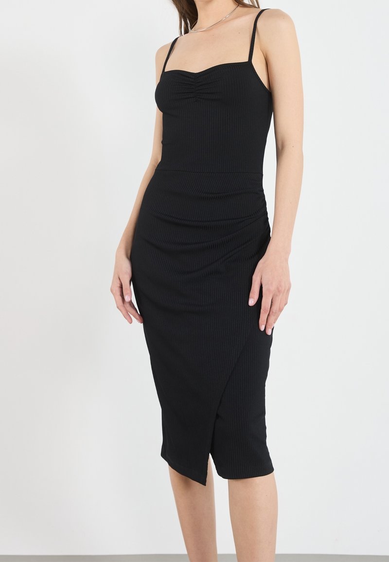 Robe noire côtelée avec un corsage ajusté, des bretelles spaghetti réglables et une jupe froncée avec une fente latérale, fabriquée en tissu doux.