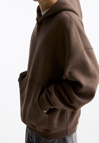 Sweat-shirt à capuche marron avec une coupe décontractée, poche avant kangourou et poignets côtelés. Le tissu semble doux avec une texture lisse.