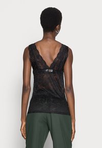 Donna con i capelli corti che indossa un top senza maniche in pizzo nero con una profonda scollatura a V sul retro e pantaloni verdi scuri, in piedi contro uno sfondo chiaro e semplice.