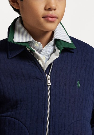 Jūras spēku zilu, quiltētu jaku ar zaļām apkakles detaļām, kas aprīkota ar sudraba rāvējslēdzēju un izšūtu logotipu. Zem tās valkāta gaiši zila svītraina krekla ar baltām akcentiem.