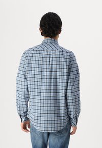 Man with dark curly hair wearing a long-sleeve blue plaid shirt and blue jeans, standing facing away against a white background.