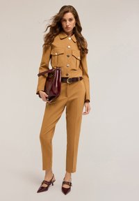 Jumpsuit su misura color tan con dettagli in corrispondenza dei bottoni, due tasche sul petto e cintura abbinata. Il modello tiene una borsa bordeaux e indossa scarpe con tacco bordeaux.