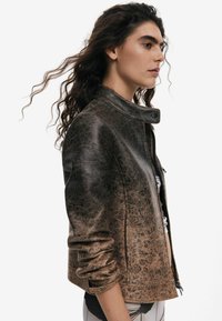 Veste en cuir marron avec un motif texturé subtil, fermeture éclair et col montant. Présente une silhouette ajustée et des manches retroussées.