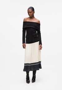 Black off-the-shoulder blazer with two front pockets, paired with a pleated cream skirt featuring a navy ruffle and a decorative waistband.