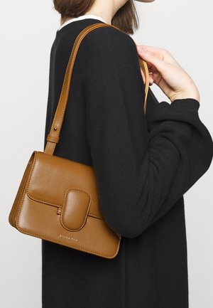 Hnědá kožená kabelka obdélníkového tvaru s předním klopovým zapínáním a dlouhým nastavitelným popruhem, vystavená na černém svetru.