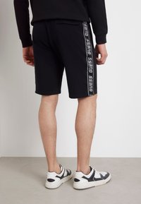 Pantalones cortos negros con una textura suave, presentan un diseño lateral etiquetado como "GUESS" en gris. Llevados con zapatillas negras y blancas.