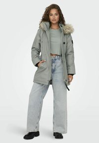 Parka grise avec capuche en fausse fourrure, fermetures éclair et boutons-pression, poches avant et accents noirs. Portée par-dessus un crop top vert et un jean en denim clair.