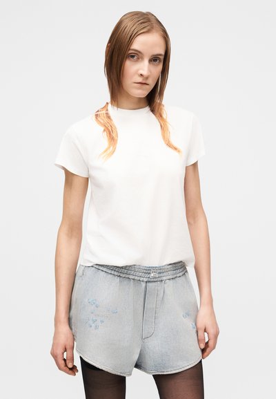 Jovem com cabelo castanho claro liso veste uma t-shirt branca, calções de ganga claros e collants pretos contra um fundo branco simples.