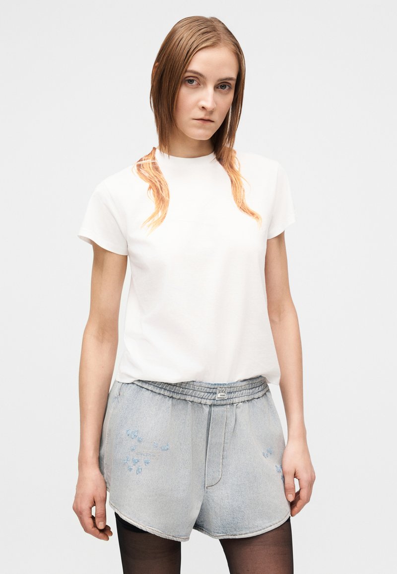 Ung kvinde med glat lyst brunt hår iført en hvid t-shirt, lyse denimshorts og sorte strømpebukser mod en ensfarvet hvid baggrund.