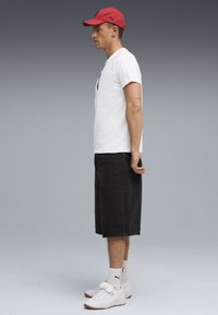 Rote Mütze, weißes T-Shirt mit Logo, schwarze knielange Shorts, weiße Sneakers mit Akzent; legere Outfit mit einem konzentrierten Profil.