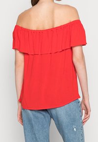 Top rosso scollato sulle spalle con un design a strati di rouches, caratterizzato da una texture leggera e un orlo leggermente curvo, abbinato a jeans blu comodi.