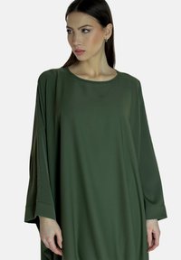 Abito lungo e morbido in tessuto verde scuro, con scollo rotondo e fessure laterali alle maniche per una maggiore libertà di movimento.