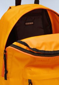 Mochila de tela naranja con acentos negros, que presenta una apertura grande, un compartimento con cremallera y una etiqueta de logo en el interior. Material texturizado.