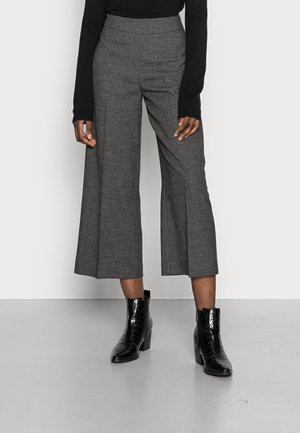 Personne portant un pantalon gris large et court, un haut noir à manches longues et des bottines noires à talons, debout sur un sol gris.