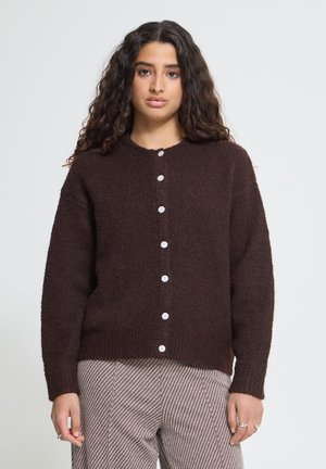 Jeune femme aux longs cheveux bouclés portant un cardigan marron foncé boutonné et un pantalon clair rayé, debout devant un fond clair uni.