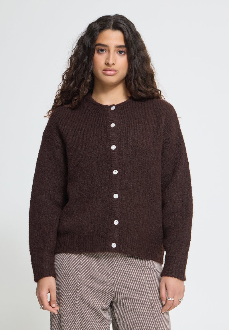 Jeune femme aux longs cheveux bouclés portant un cardigan marron foncé boutonné et un pantalon clair rayé, debout devant un fond clair uni.