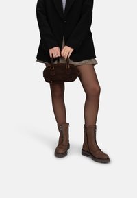 Bottines en suede brun avec fermetures éclair, associées à un blazer noir, une jupe à motifs et un petit sac à main brun. Texture lisse et design robuste.