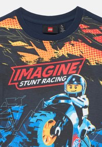 Schwarzes T-Shirt mit einem bunten Grafikdesign eines LEGO-Motorradfahrers. Das Design enthält orangefarbene, gelbe und blaue Akzente mit dem Slogan "IMAGINE STUNT RACING".