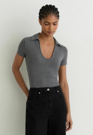Junge Frau mit geflochtenem Haar, die ein graues, kurzärmliges Oberteil mit Kragen und schwarze, hoch taillierte Hose vor einer schlichten Wand trägt.