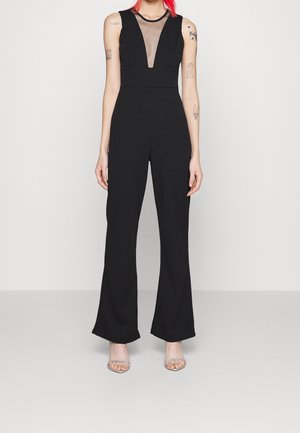 Vrouw met rood haar die een mouwloze zwarte jumpsuit draagt met een transparante V-hals en zilverkleurige open-toe hakken, staand tegen een effen achtergrond.