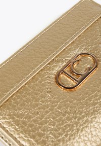 Guldig texturerat läderkortfodral med en framträdande cirkulär logotypdetalj. Slät yta med präglade detaljer och kontrasterande sömnad.