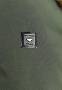 Tissu vert foncé à texture lisse et à légère brillance, avec un patch rectangulaire en cuir noir embossé du nom de la marque et du logo.