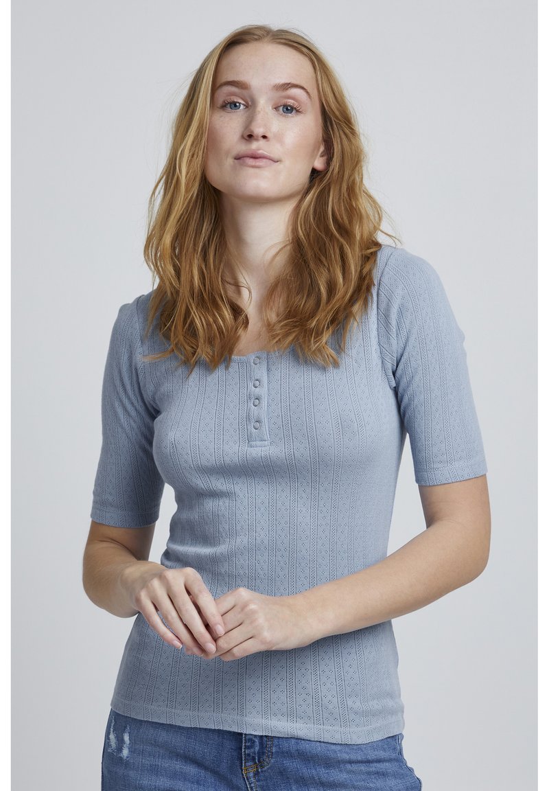 Gaiši zils ribots tops ar īsām piedurknēm, apļveida kakla izgriezums un piecu pogu aizdare. Audumam ir struktūrizēts raksts.