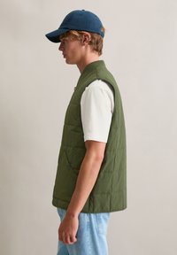 Bezrekawnik puchowej kamizelki w kolorze oliwkowej zieleni z pikowaną teksturą, z zaokrąglonym kołnierzem, przednimi kieszeniami i zapięciem na guziki. Nosi się ją na białej koszuli.