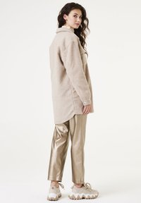 Camisa chaqueta de forro polar beige con cuello, bolsillos laterales y un corte suelto, combinada con pantalones dorados brillantes ajustados y zapatillas gruesas.