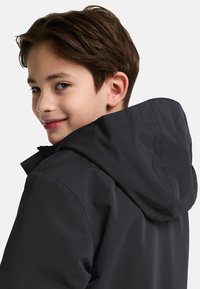 Giacca nera impermeabile con cappuccio, caratterizzata da una texture liscia e un design semplice, indossata da un giovane con capelli corti castani e un leggero sorriso.