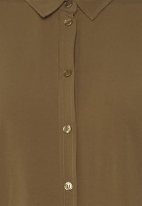 Bruine blouse van gladde stof met een klassieke kraag, voorzien van zes ronde knopen en subtiele stiksels langs de knoopsluiting.