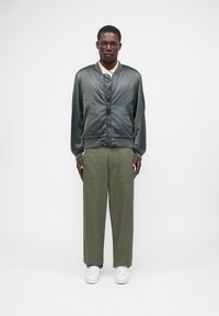 Casaco bomber masculino em satin cinza-escuro com frente acolchoada, combinado com calças largas verde-oliva e ténis brancos.