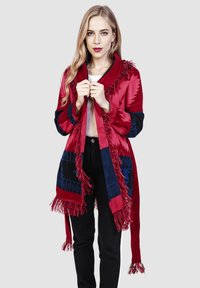 Chal rojo con patrones geométricos azules, bordes flecados y un amplio cuello, hecho de una tela suave, combinado con pantalones negros.