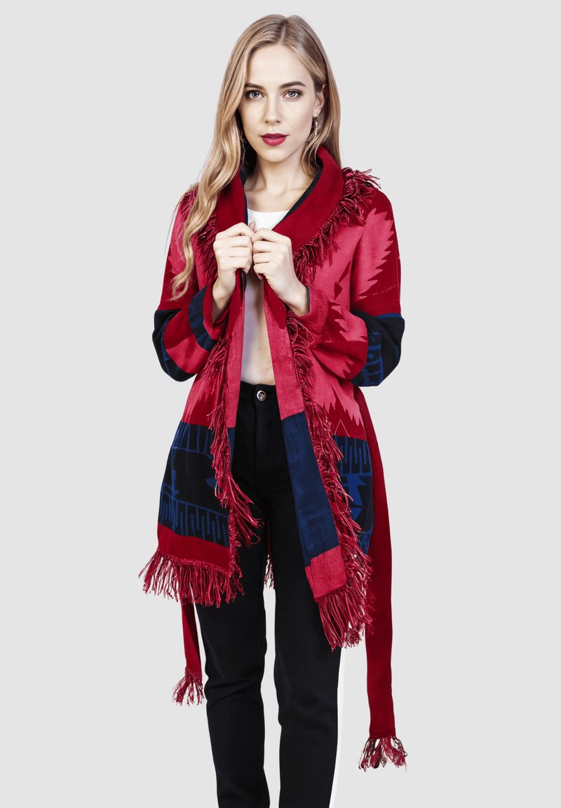 Chal rojo con patrones geométricos azules, bordes flecados y un amplio cuello, hecho de una tela suave, combinado con pantalones negros.