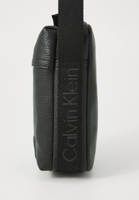 Black textured leather bag with a rectangular shape. Features a wide strap adorned with "Calvin Klein" in embossed lettering.