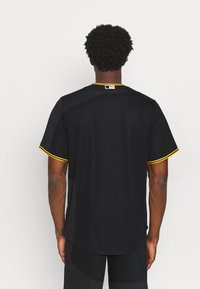 Uomo di spalle che indossa una maglietta sportiva nera a maniche corte con rifiniture gialle e un logo bianco sul colletto posteriore.
