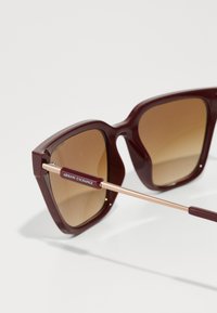 Ochelari de soare colorați burgundy, supradimensionați, cu lentile din gradient maro. Prezintă brațe din metal cu nuanță de aur, cu branding "Armani Exchange" pe tâmple.