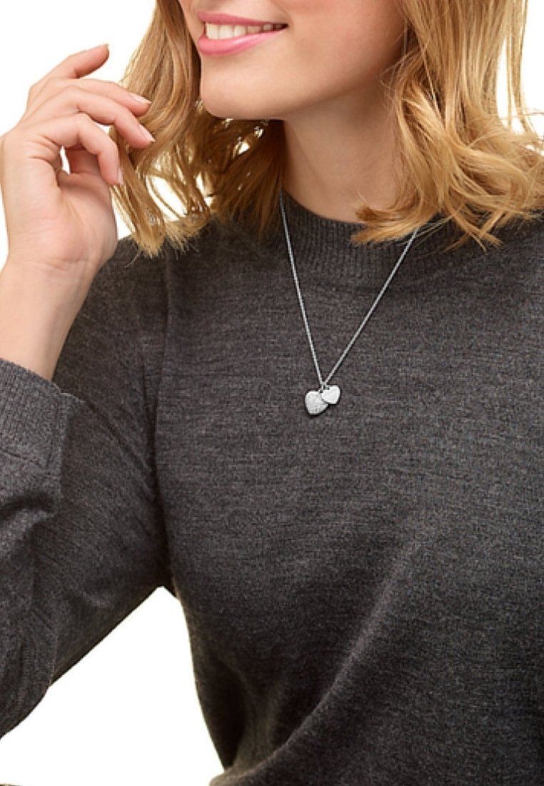 Femme portant un pull gris foncé et un collier en argent avec deux pendentifs en forme de cœur, souriante et touchant ses cheveux blonds.