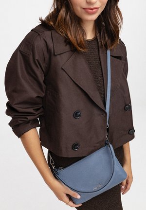 Femme portant une veste courte marron foncé et une robe en maille, tenant un sac bandoulière bleu à surface texturée et avec logo.