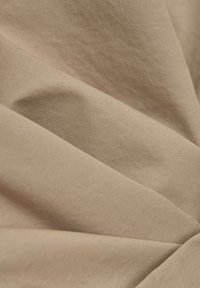 Béžová tkanina s hladkou strukturou, se zavěšenými záhyby, které vytvářejí hloubku a jemné stíny, zdůrazňující měkkost materiálu.