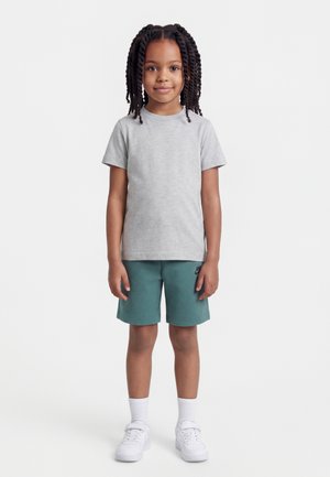 Niño con cabello trenzado que lleva una camiseta de manga corta gris claro, pantalones cortos verde azulado, calcetines blancos y zapatillas blancas, de pie sobre un fondo blanco.