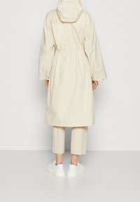 Impermeabile beige chiaro con cappuccio, vita elastica e maniche lunghe. Abbinato a pantaloni cropped coordinati e sneakers bianche. Texture liscia.