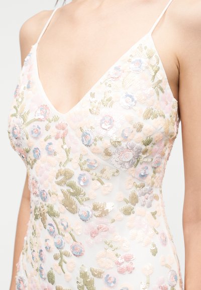 Γυναίκα που φοράει λευκό φόρεμα με λεπτές τιράντες, βαθύ V λαιμόκοψη και παστέλ κεντήματα με παγιέτες σε floral σχέδιο.